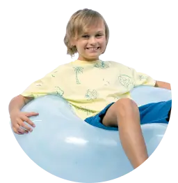 Chłopiec w jasnej koszulce siedzi na niebieskiej piłce i uśmiecha się